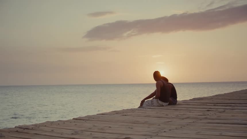 A silhouetted couple enjoys a peaceful sunset while sitting on a wooden pier over the ocean as a swimmer plays nearby