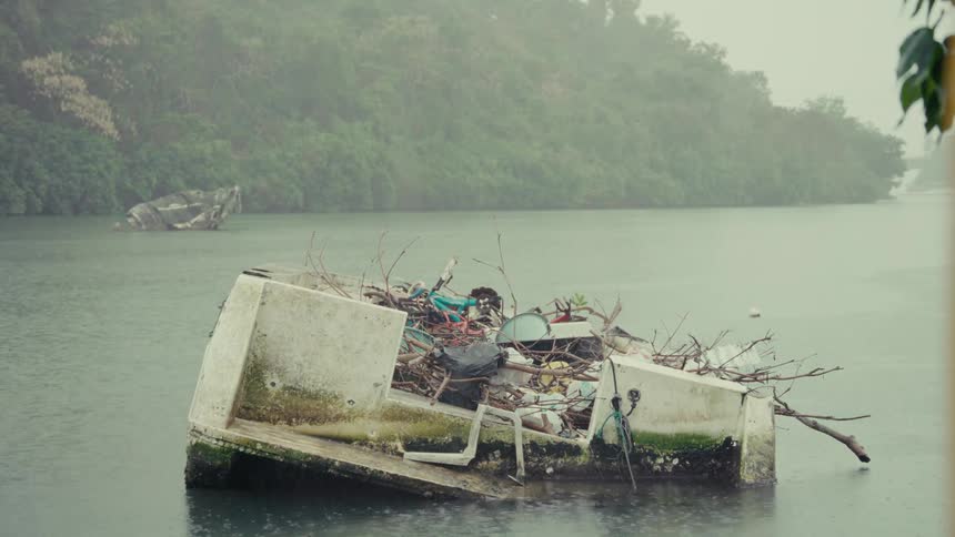 Rain falls on a half-sunken boat filled with garbage, highlighting water pollution in a tropical river