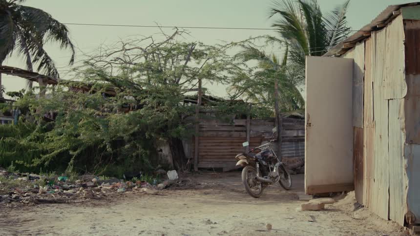A desolate and impoverished shantytown scene featuring a parked motorcycle next to a rusty tin shack