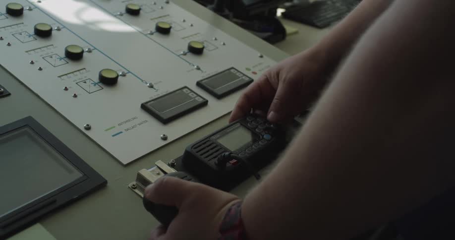 A crew member's hands operate a VHF radio at the control panel on a ship's bridge