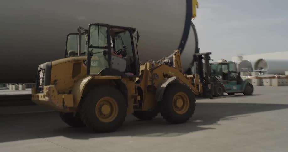A construction worker drives a yellow front-end loader, towing a massive wind turbine tower section across an industrial yard
