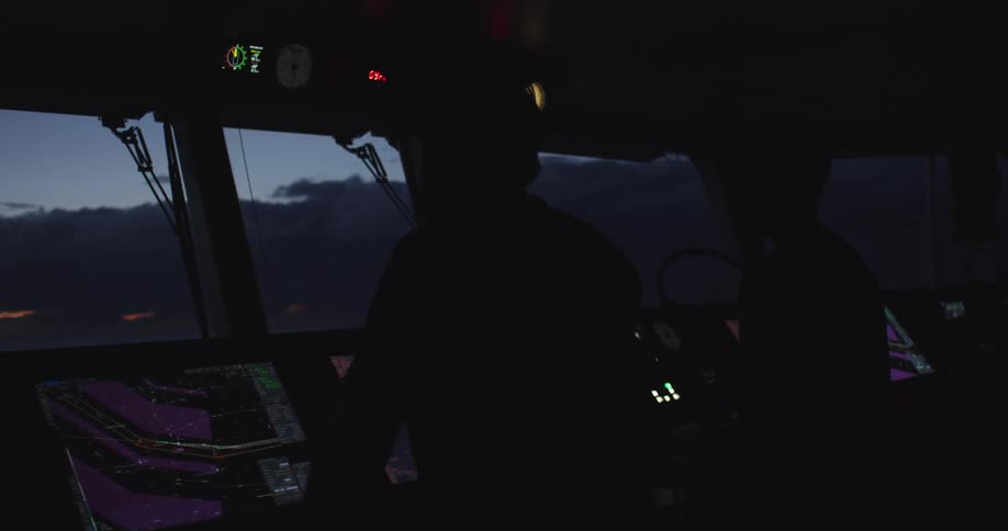 The silhouettes of two sailors stand on the darkened bridge of a ship, navigating the ocean at twilight using illuminated control panels and screens