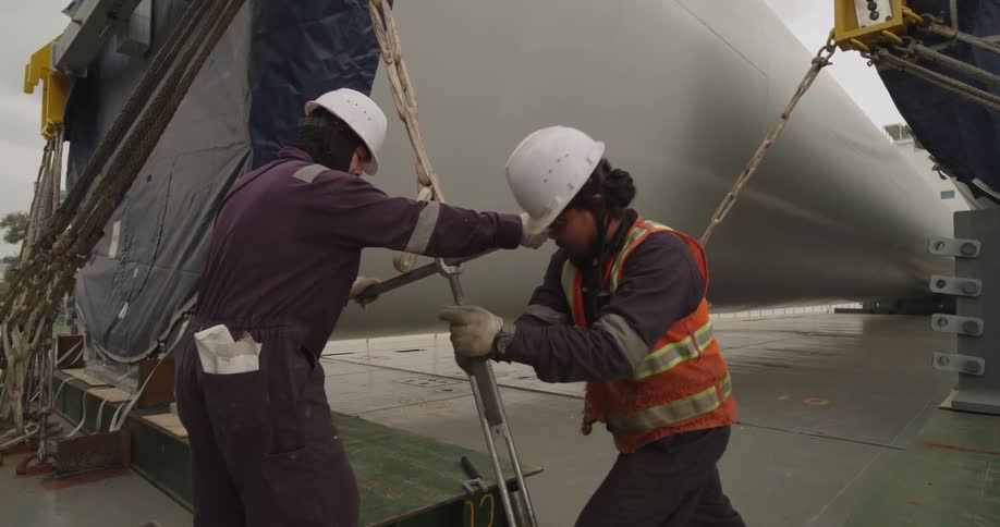 Two industrial workers in hard hats use a tool to tighten the lashings securing a large wind turbine tower to the deck of a cargo ship