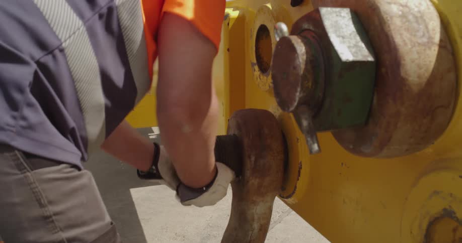 A worker wearing safety gloves unscrews a large rusty nut from a piece of heavy yellow industrial machinery