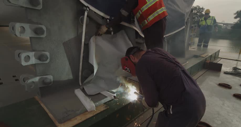 A welder in a protective helmet kneels to weld a large metal structure on a construction site, with sparks flying