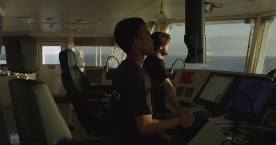 Two crew members on a research vessel navigate the ship from the bridge while looking out at the open ocean