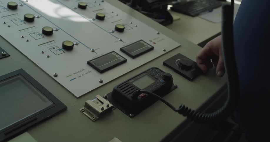 A person on a ship's bridge uses the radio and then turns a dial on the control panel
