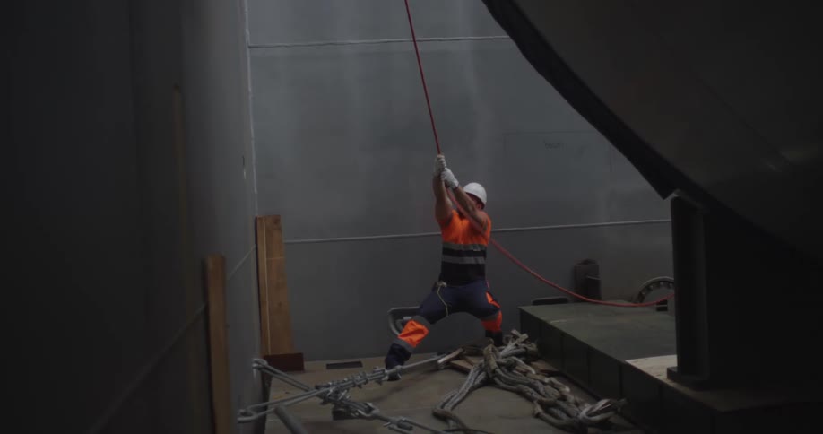 A construction worker in a hard hat and safety vest strains as he pulls on a red rope inside a large industrial building