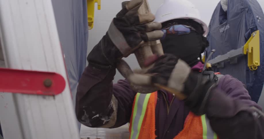 A construction worker wearing safety gear carefully attaches a large metal shackle to a heavy-duty lifting strap