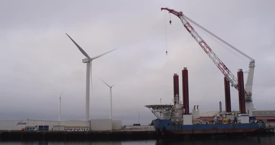 A massive crane vessel for wind turbine installation is docked in an industrial port with onshore wind turbines spinning