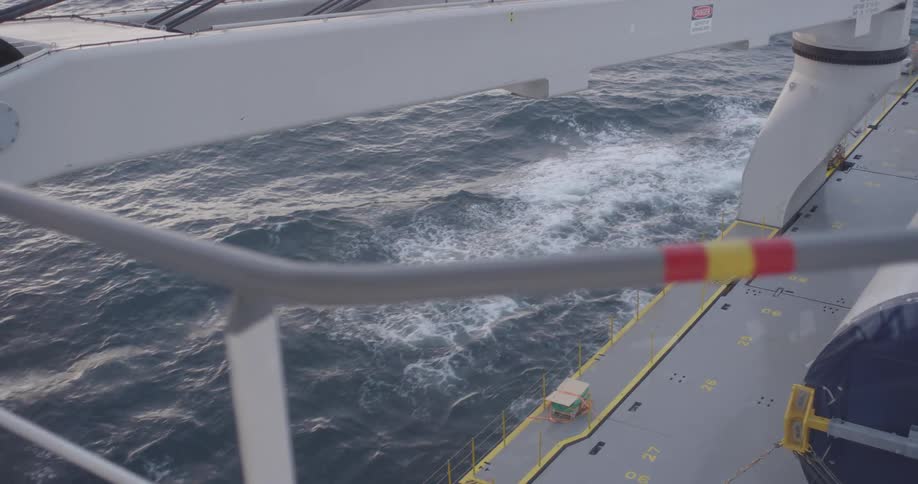 A high-angle view from a large ship's deck shows the vessel moving through the choppy ocean water