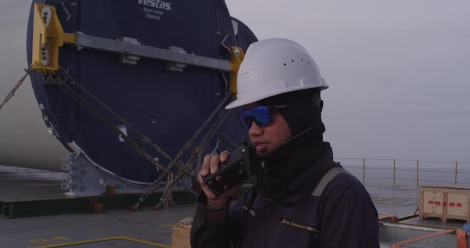 An offshore construction worker wearing a hard hat and safety glasses communicates via walkie-talkie on the deck of a transport ship at sea