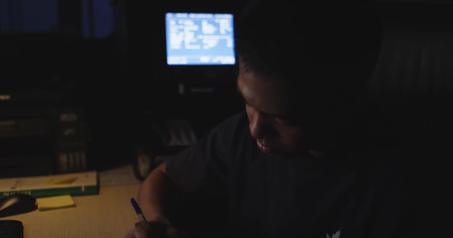 A focused young man studies at his desk in a dark room in slow motion, illuminated by a single lamp