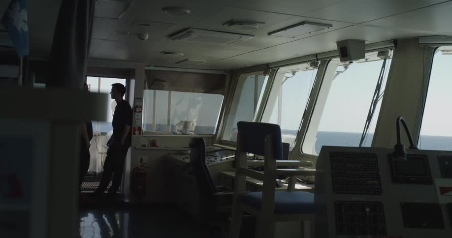 Two crew members have a conversation on the dimly lit bridge of a ship while sailing on the open ocean