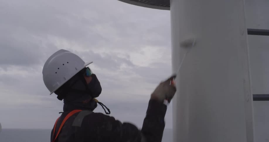 A worker in full safety gear carefully paints a large white mast on a ship out at sea