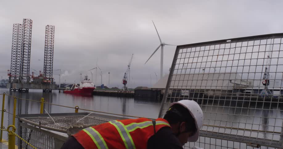 A dock worker handles heavy rigging in an industrial port with offshore rigs and wind turbines in the background