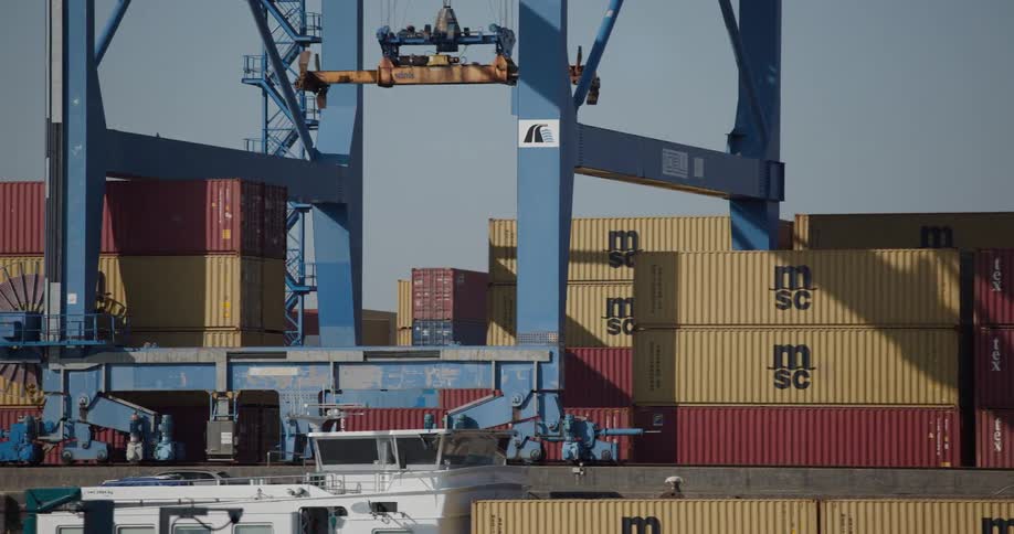 A large blue gantry crane moves along its tracks above stacks of shipping containers at a busy port terminal