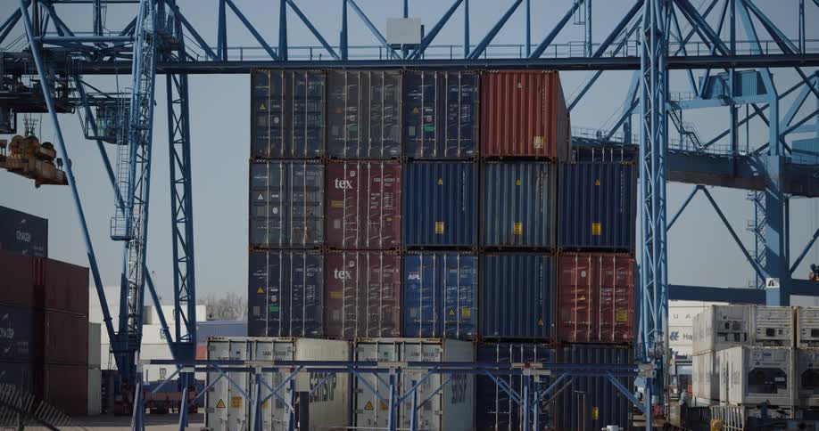 A tall stack of colorful shipping containers sits under a large gantry crane at an industrial port
