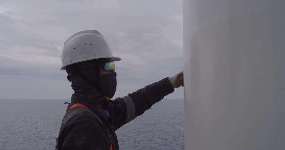 A maritime worker in full safety gear carefully paints a white column on a ship at sea under an overcast sky