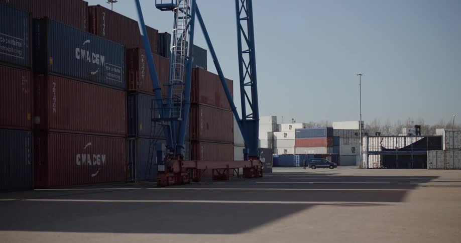 A person exits their car in a vast industrial shipping yard filled with stacked containers and a large crane