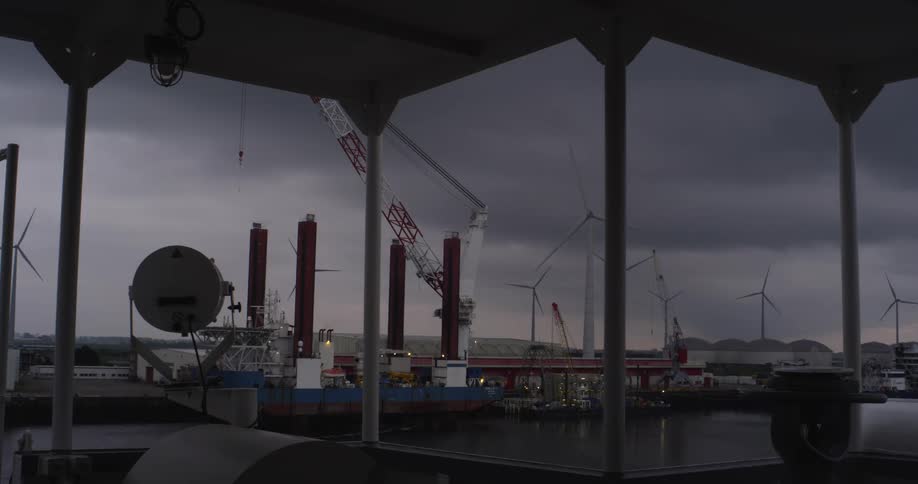 An industrial harbor with a massive jack-up rig and wind turbines stands under a brooding, overcast sky at dusk