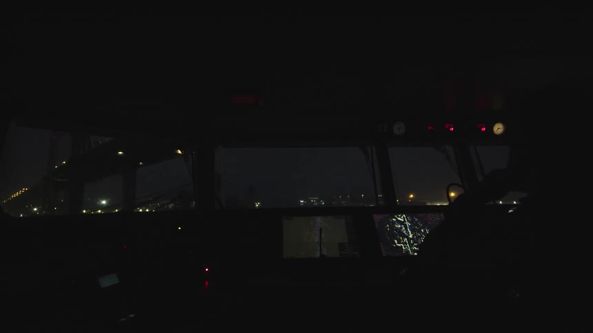 Inside a ship's dark bridge at night, with a view of the illuminated port and a silhouette of the captain