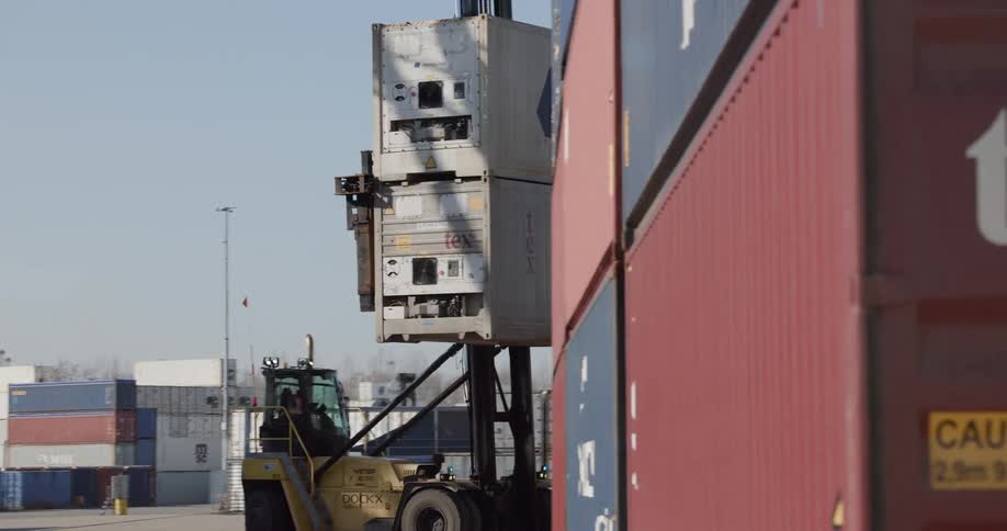 A large yellow reach stacker transports two stacked shipping containers and places them beside another stack at a bustling port