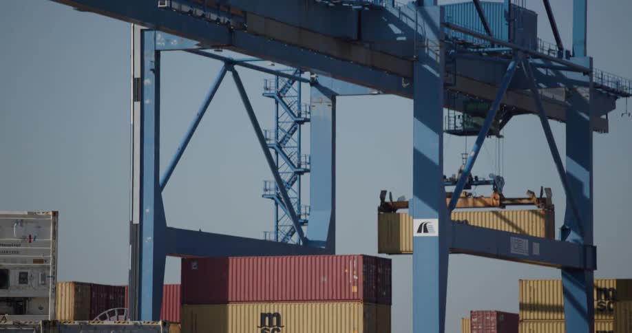 A large blue industrial port crane lifts a yellow shipping container from a barge as the camera tilts up