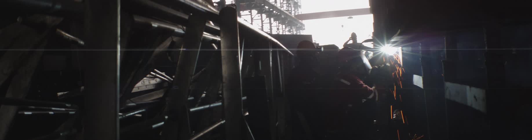 A welder in protective gear works on a metal structure in a vast, sunlit industrial factory as sparks fly