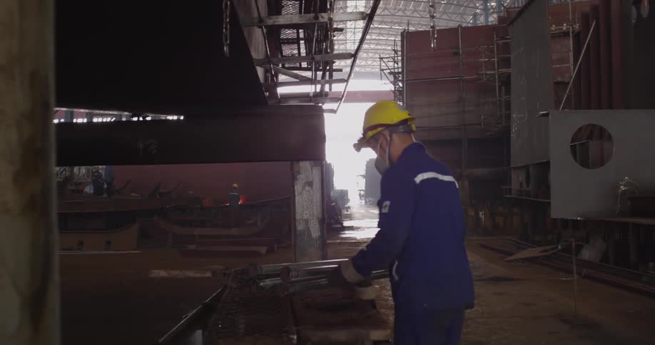 Shipyard worker in full safety gear meticulously cleans metal rods inside a massive hangar