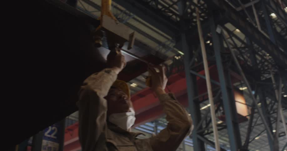 A factory worker in a hard hat and mask meticulously inspects a large steel component suspended by a crane