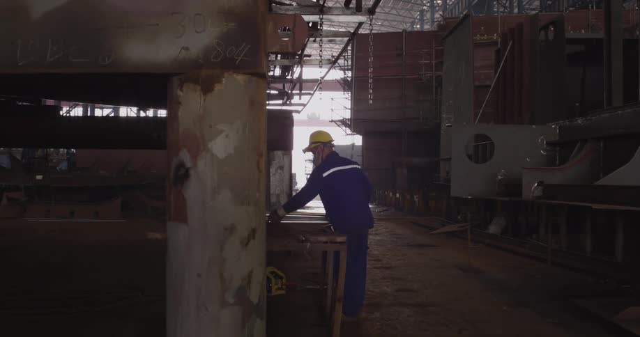 A shipyard worker in protective gear carefully grinds a metal plate in a large, dark industrial workshop