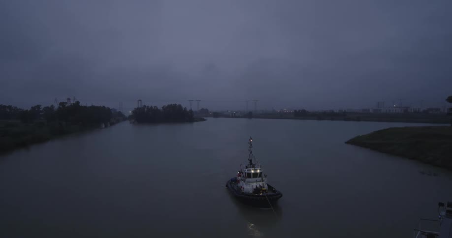 A lone tugboat with its lights on floats in a wide, misty river on a dark and foggy evening