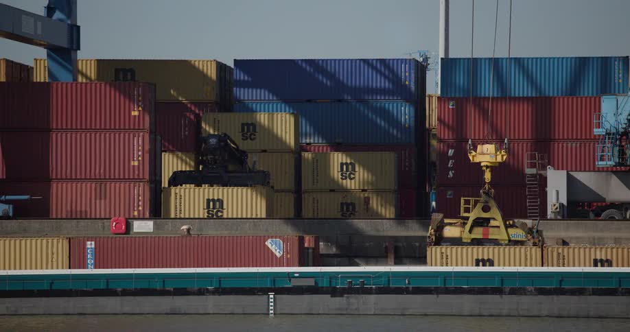 A large yellow port crane slowly unloads shipping containers from a barge at a bustling industrial harbor