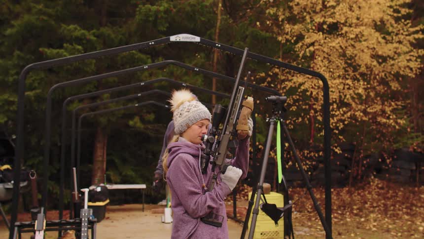A young girl in a winter hat inspects a high-powered rifle at an outdoor shooting range in the autumn