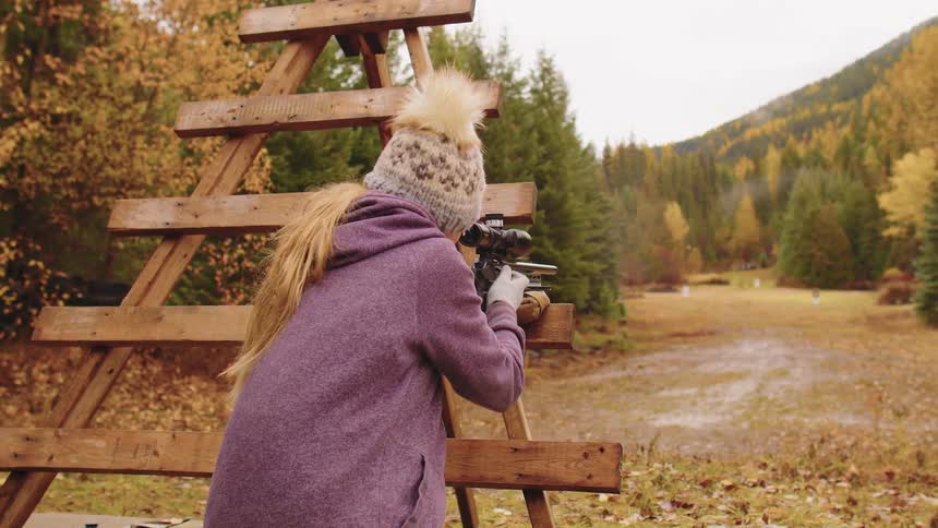 A female shooter fires a rifle in slow motion at an outdoor shooting range surrounded by autumn forests