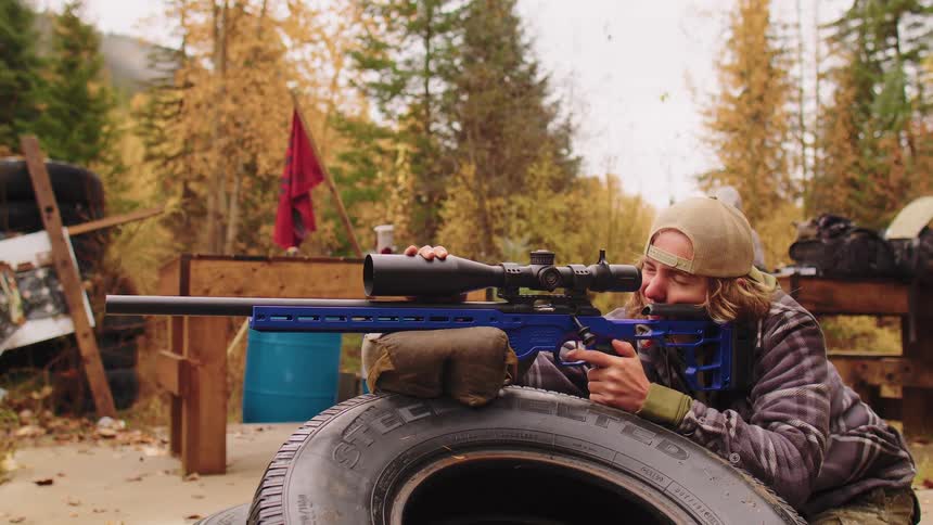 A young marksman with long hair aims and operates the bolt action on a blue sniper rifle at a rural outdoor range in autumn