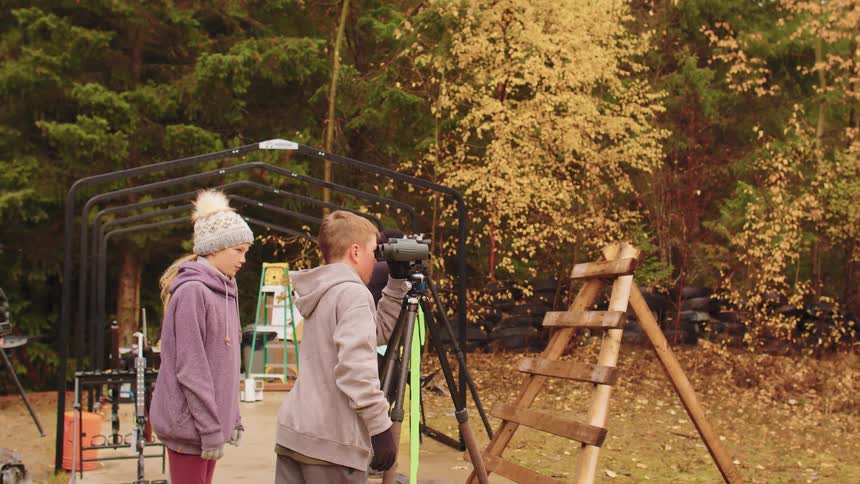 Two children take turns looking through a spotting scope at an outdoor range on a beautiful autumn day