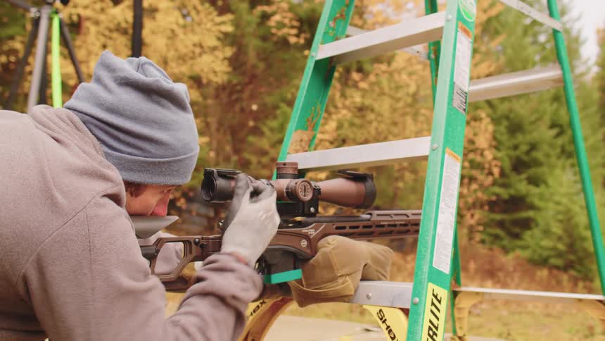 A competitive shooter carefully aims and fires a scoped rifle stabilized on a ladder in an autumn forest