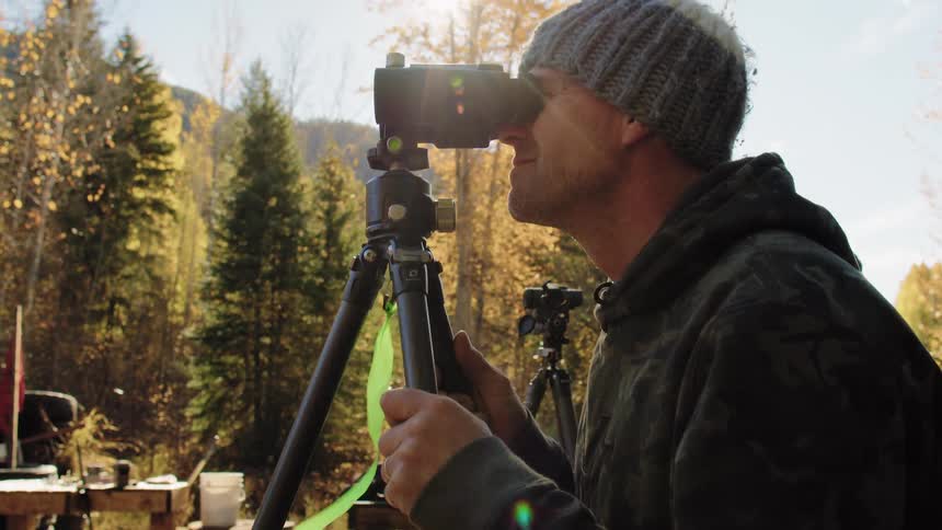 Outdoorsman in a beanie and camouflage hoodie scans the autumn forest through a spotting scope with the sun flaring in the background