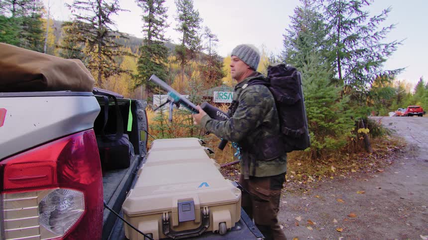 A man in hunting gear with a backpack carefully assembles and inspects his rifle from the back of his truck