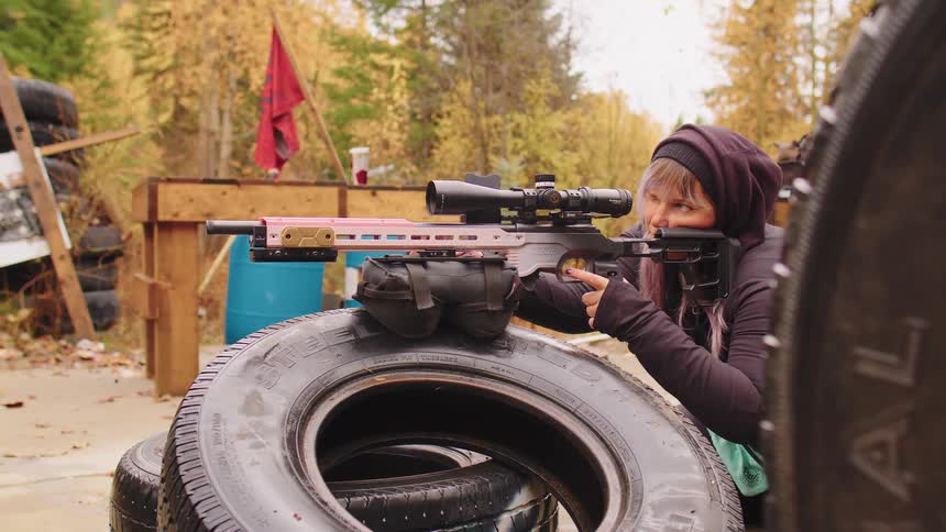 A woman in a hoodie lines up her shot with a sniper rifle resting on tires at an outdoor practice range during autumn