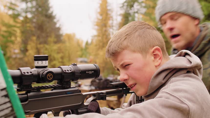 A young boy squints as he takes aim with a scoped rifle while his father instructs him in the background