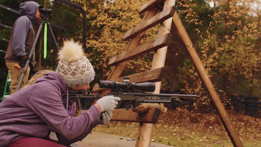 A female shooter aims and fires a bolt-action sniper rifle from a wooden barricade at an outdoor range while a spotter watches in the background