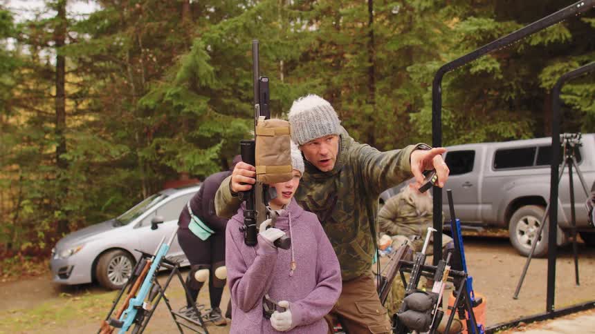 An instructor guides a young girl holding a large rifle and points toward a target during an outdoor shooting lesson