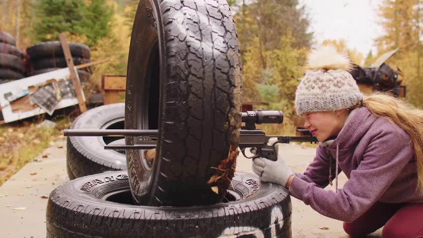 A young woman wearing a winter hat practices shooting a scoped pistol in slow motion at an outdoor range, resting the gun on old tires