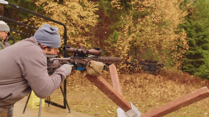 A man in a beanie fires a scoped rifle from a barricade at an outdoor shooting range in slow motion
