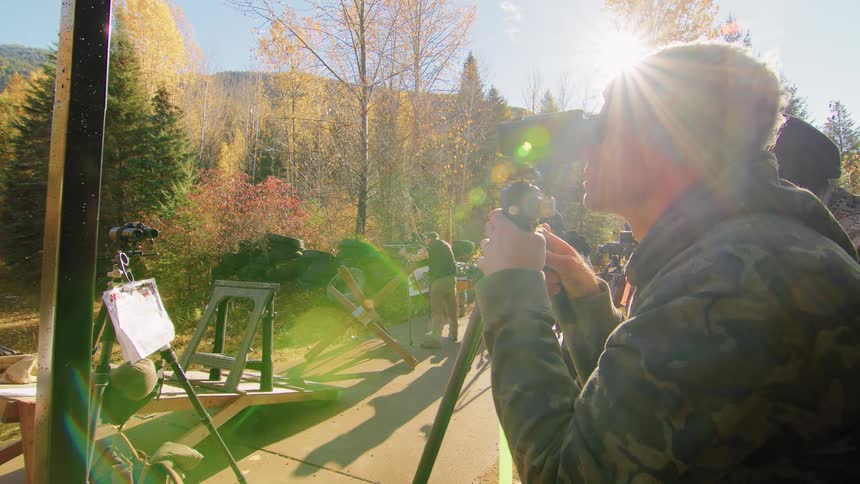 A man in camouflage uses a spotting scope to observe an outdoor shooting range on a sunny autumn day
