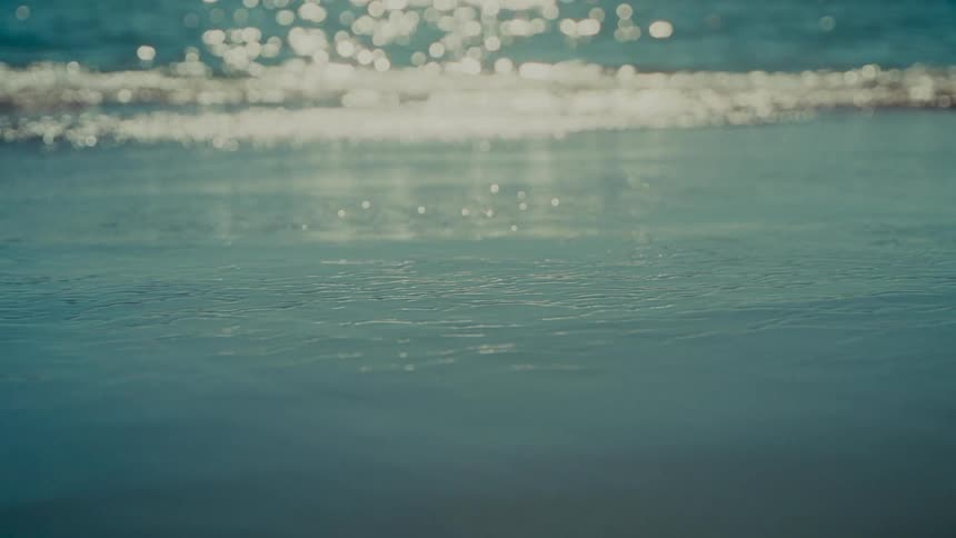 A tranquil close-up shot captures the calm ocean water lapping at the shore, with the sun creating a sparkling bokeh effect