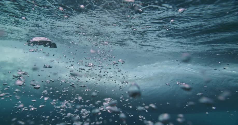 A slow-motion underwater shot captures a wave passing overhead, creating a beautiful chaos of bubbles that rise towards the split-level view of the sky